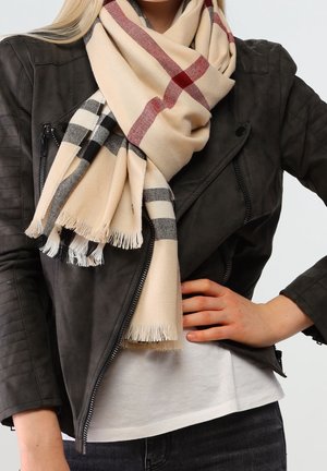 Personne blonde portant une veste en cuir noire et une écharpe beige à motifs écossais rouges et noirs, la main reposant sur la hanche.