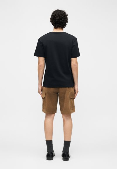 Άνδρας με σγουρά μαλλιά, φοράει μαύρο κοντομάνικο μπλουζάκι, καφέ cargo σορτς, γκρι κάλτσες και μαύρα παπούτσια, στέκεται γυρισμένος προς τα πίσω.