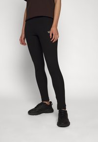 Černé legíny vyrobené z pružného materiálu, s úzkým střihem a vyšitým logem. Nosené s černými sportovními botami na neutrálním pozadí.