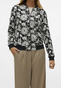 Kurtka bomber w kwiaty w kolorze czarnym z białym nadrukiem kwiatowym, z pełnym zapięciem na zamek i ściągaczami przy mankietach. Połączona z beżowymi spodniami, podkreślającymi luźny krój.