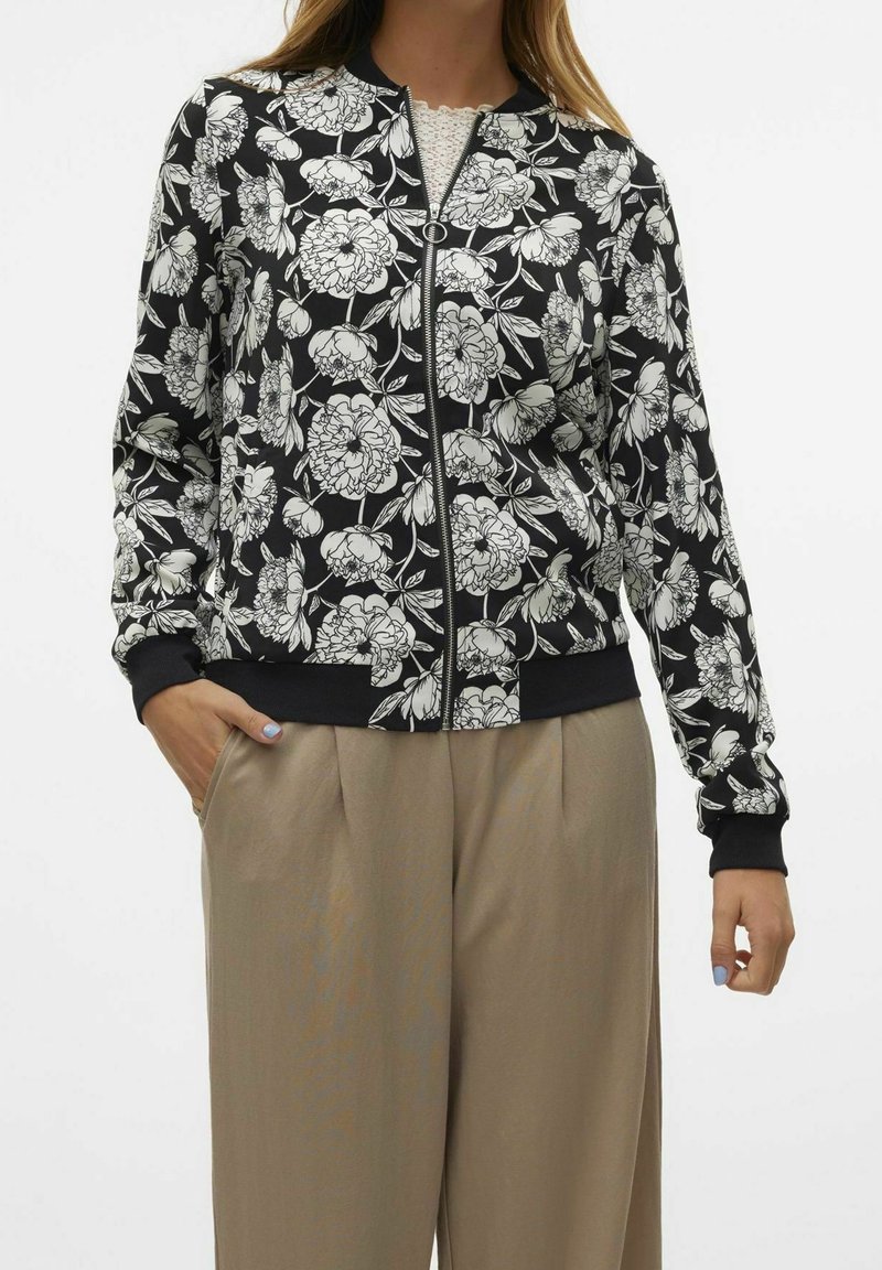 Kurtka bomber w kwiaty w kolorze czarnym z białym nadrukiem kwiatowym, z pełnym zapięciem na zamek i ściągaczami przy mankietach. Połączona z beżowymi spodniami, podkreślającymi luźny krój.