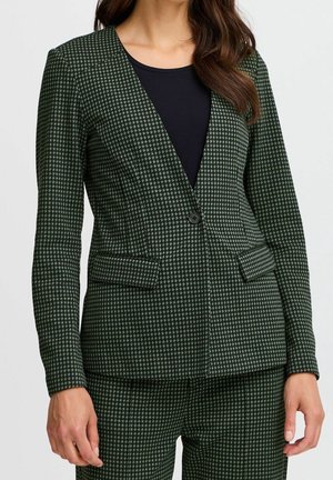 Kvinde iført blazer og matchende bukser med mørkegrønt og sort hundetandsmønster samt en sort top indenunder.