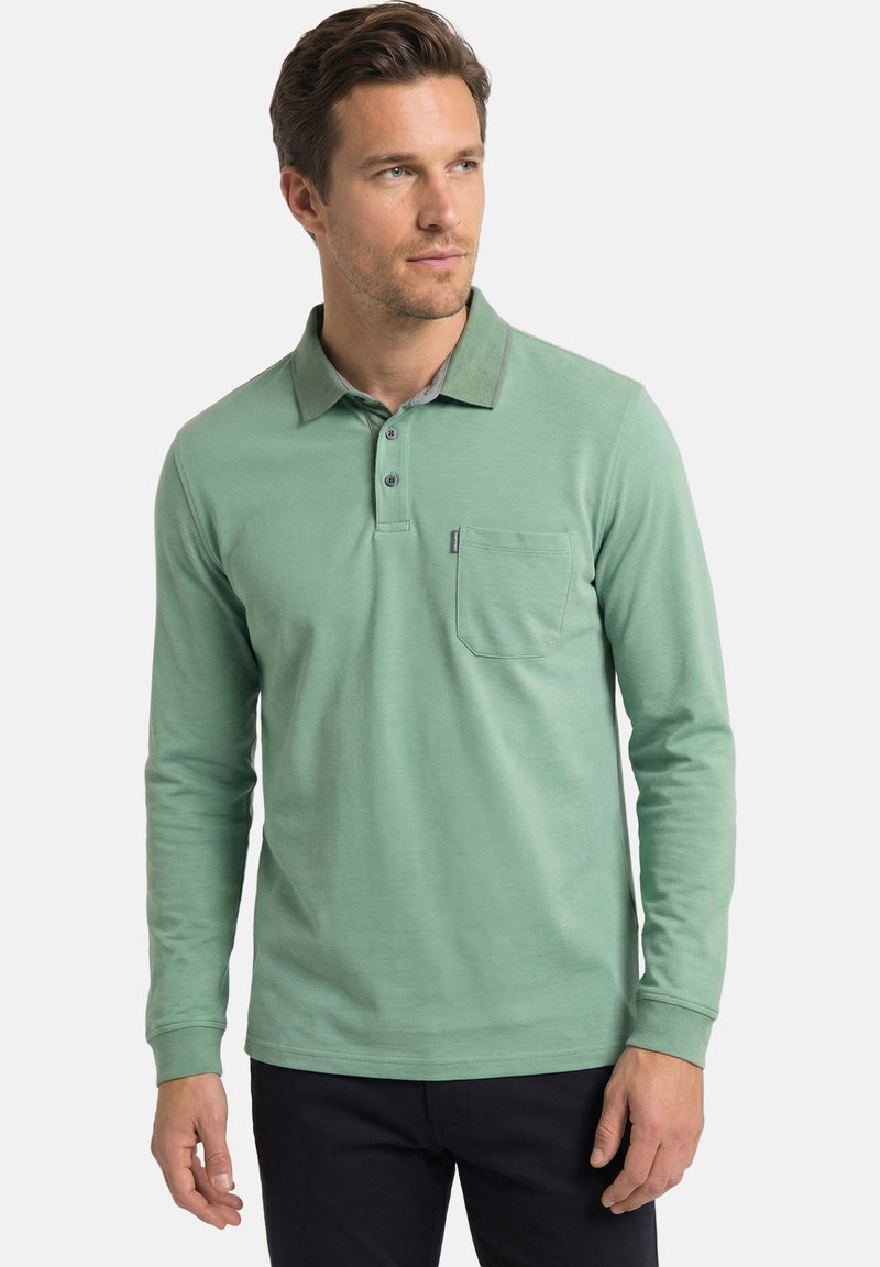 Homme portant un polo vert à manches longues avec une poche poitrine, debout devant un fond clair uni, regardant légèrement vers la droite.