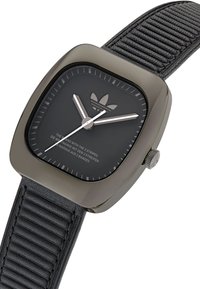 Reloj negro en forma de cuadrante con una correa de silicona negra texturizada. Presenta agujas plateadas para las horas y los minutos, y un logo en relieve en la esfera.