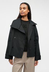 Abrigo negro tipo trench con cuello, botones de doble botonadura y diseño a medida. Lleva puesto un suéter negro con un patrón geométrico.