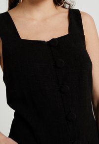 Black linen dress featuring a square neckline, thick straps, and large round button closures down the front. Simple and textured fabric.