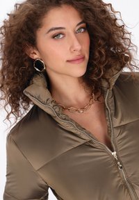 Wypchana khaki kurtka z wysokim kołnierzem i zamkiem błyskawicznym z przodu. Złoty naszyjnik łańcuszkowy i ośmiokątne kolczyki dopełniają stylizację. Widoczne kręcone włosy.