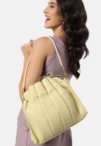Gul läderhandväska med plisserad design, guldhardware och ett enda axelband. Slät textur och medelstorlek.