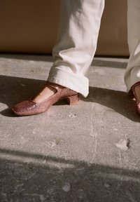 Różowe, brokatowe buty Mary Jane na niskim, blokowym obcasie, z kwadratowym noskiem i paskiem wokół kostki, w połączeniu z białymi, prostymi spodniami na teksturowanej podłodze.