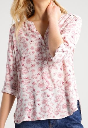 Kvinde iført en hvid bluse med lyserøde blomsterprint og op rullede ærmer, kombineret med blå jeans, hånd rører ved nakken.