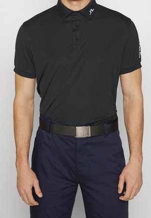 Mężczyzna w czarnej koszulce polo z krótkim rękawem, granatowych spodniach i czarnym pasku z metalową klamrą z logo Under Armour.