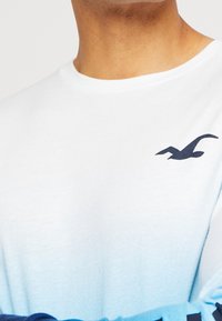 Vit t-shirt med kort ärm och gradientdesign i blått, med en marinblå logotyp av en fågel på bröstet. Tillverkad i bomull, avslappnad passform.