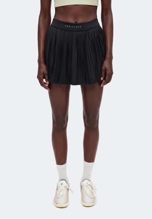 Vrouw die een zwart geplooid tennisrokje draagt met "ENDLESS" op de tailleband, een beige cropped top, witte sokken en witte sneakers.