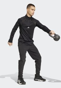 Zwarte sportieve pullover met ritssluiting en logo, gecombineerd met zwarte taps toelopende broek en zwarte sneakers, terwijl hij een kettlebell vasthoudt in een workouthouding.