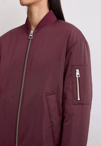 Bordeaux bomberjack met een geribde kraag, zilveren ritssluiting en een ritszak op de linker mouw. Glad, matte textuur.