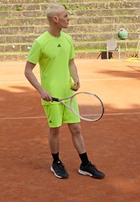 Camiseta y pantalones cortos deportivos de color verde neón, zapatillas de tenis negras, sosteniendo una raqueta de tenis y una pelota, en una cancha de tierra batida con escaleras de piedra al fondo.