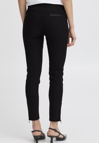 Žena u uskim crnim hlačama s pozadinskim džepovima zatvorenim zatvaračem i crnim niskim potpeticama s petom, stoji nasuprot obične bijele pozadine.