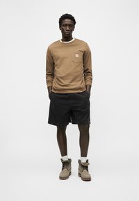 Άνδρας που στέκεται με τα χέρια στις τσέπες, φοράει ανοιχτό καφέ μακρυμάνικο πουκάμισο, μαύρες βερμούδες, λευκές κάλτσες και μπεζ μπότες σε λευκό φόντο.