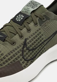 Nike Interact Run tenisice imaju teksturiranu gornju površinu od maslinasto zelene mrežice, crne naglaske, podstavljeni bijeli potplat i oznaku sa QR kodom na jeziku.