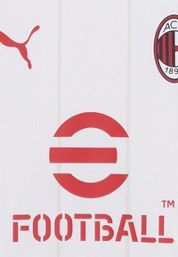 Valge jalgpallipluus, millel on punane Puma logo, punane ringikujuline disain ja tekst "FOOTBALL". Vertikaalne tekstuurne muster koos klubi embleemiga külgedel.
