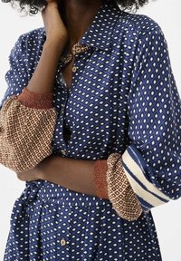 Personne portant une chemise à motifs bleus et une jupe assortie avec des poignets à motifs marron et blanc, la main reposant sur le cou et la taille.