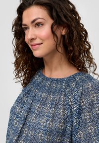Blauwe blouse met patroon, voorzien van een geplooide halslijn, florale motieven in crème en groen, en een lichtgewicht, gestructureerde stof.