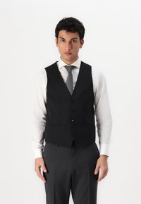 Schwarze Weste mit schmalem Schnitt, mit V-Ausschnitt, sechs Knöpfen und zwei vorderen Taschen, getragen über einem weißen Hemd und grauen Hosen.