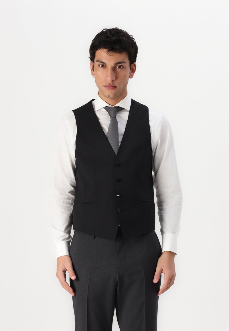 Schwarze Weste mit schmalem Schnitt, mit V-Ausschnitt, sechs Knöpfen und zwei vorderen Taschen, getragen über einem weißen Hemd und grauen Hosen.