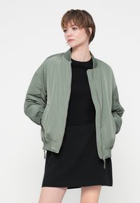 Giacca bomber verde oliva con zip frontale, polsini a coste e tasche laterali, abbinata a un vestito corto nero. Texture liscia e vestibilità rilassata.