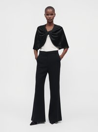 Femme debout les mains dans les poches, portant un pantalon noir à jambes larges, un haut blanc et une cape courte noire avec un détail de nœud central.