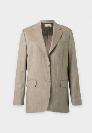 Persoon die een oversized beige krijtstreep pak draagt met broek met wijde pijpen en zwarte schoenen, staand tegen een effen witte achtergrond.