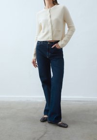 Kremfärgad stickad cardigan med rund hals och knäppning, tillsammans med mörkblå raka jeans. Modellen står på en betonggolv.