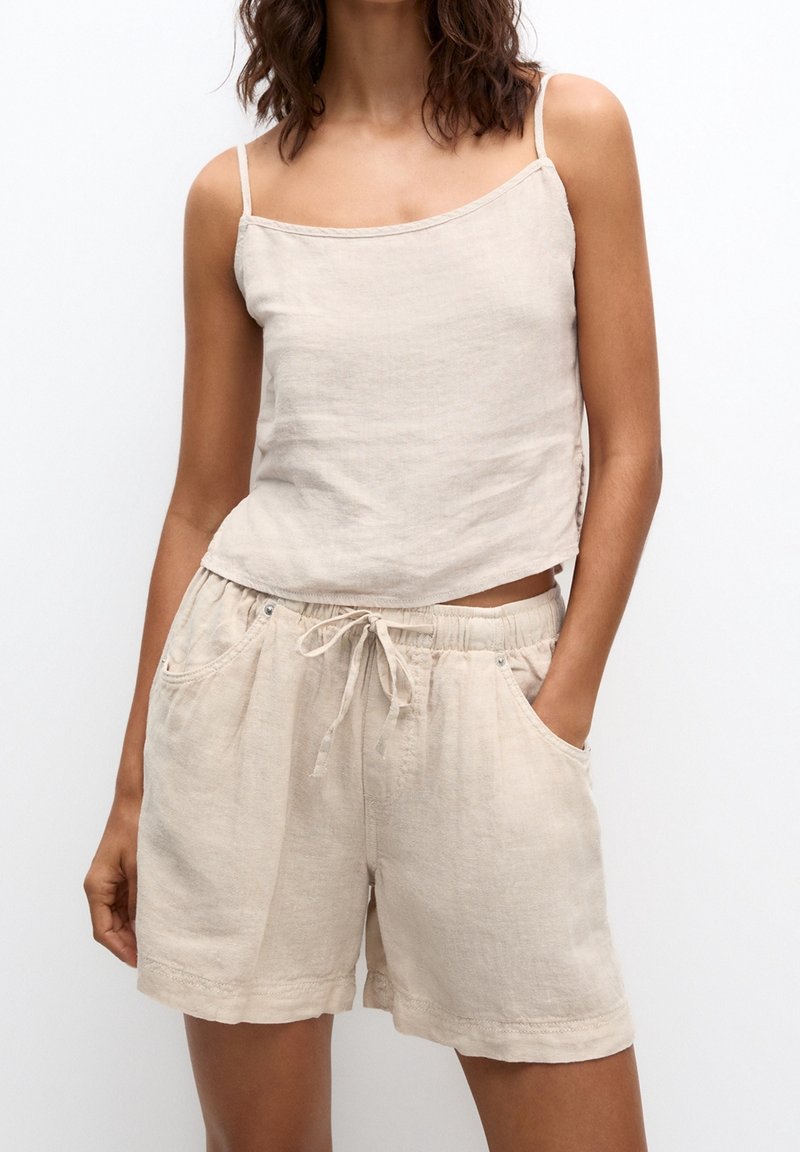 Top corto in lino beige con spalline sottili, abbinato a pantaloni corti con coulisse coordinati, dotati di due tasche frontali e una vestibilità rilassata.