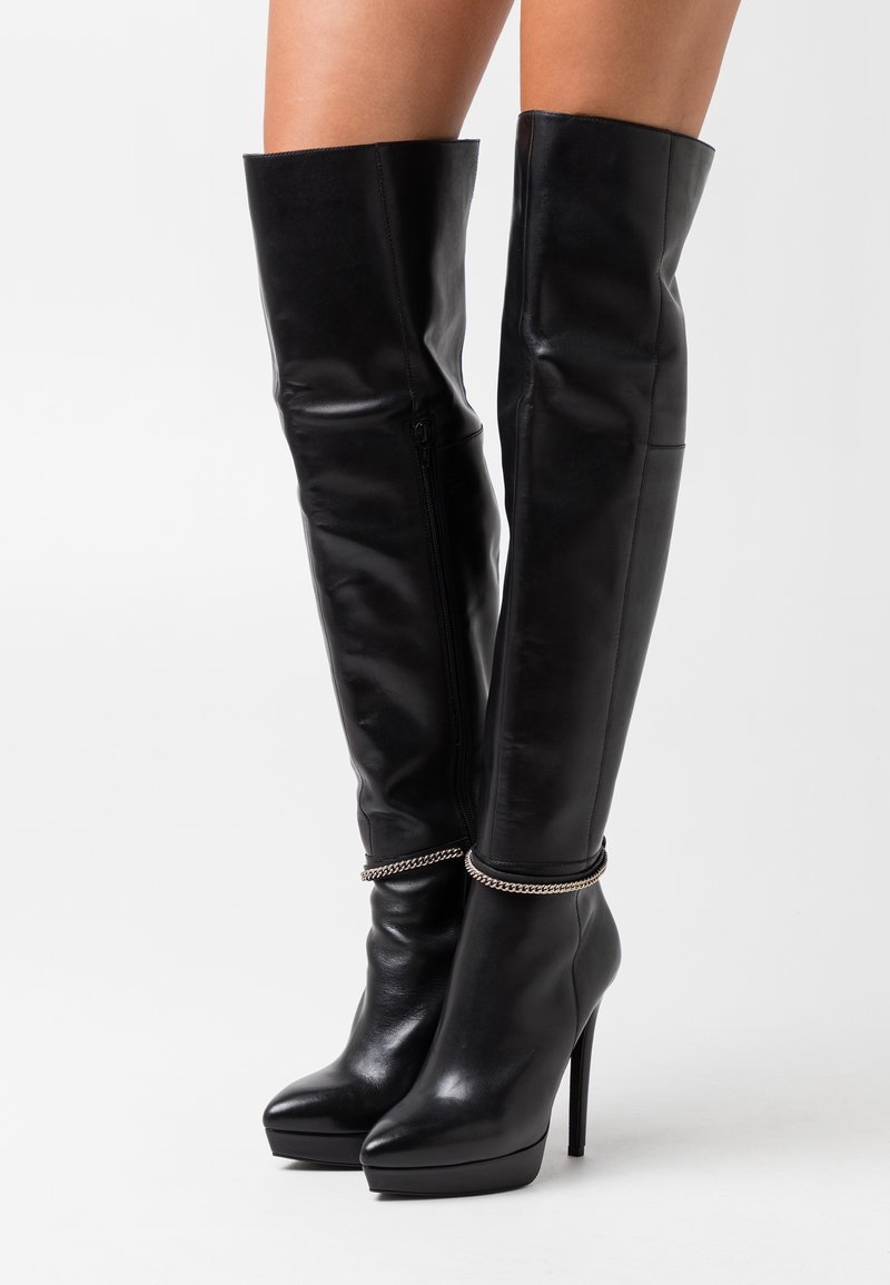 Bottes à talons aiguilles en cuir noir montant jusqu'au genou avec semelles compensées et détail de chaîne argentée portées sur des jambes fines sur fond blanc.