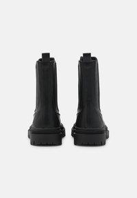 Botas de cuero negras hasta el tobillo con una textura suave, paneles laterales elásticos y suelas de goma gruesas. Cuentan con tiradores en la parte trasera.