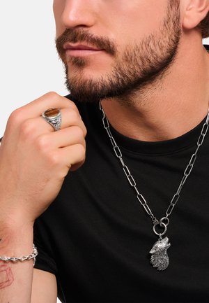 Silberner Wolf-Anhänger an einer Kettenkette; der Ring hat einen Tigerauge-Stein und wird an einer Hand getragen. Modell in einem schwarzen Shirt.