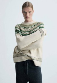 Krem pulover s uzorkom u zelenoj i smeđoj boji na yoku, rebrastim dekolteom i širokim rukavima. Uparen s tamnim rebrastim hlačama.