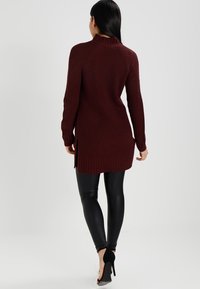 Lång burgundy ribbad stickad tröja med hög krage, sidslitningar och figurnära silhuett, ihop med tajta svarta leggings och högklackade skor.