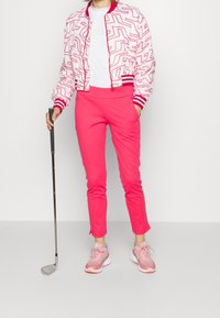 Růžová bomber bunda s červeným zigzag vzorem, bílý tričko, jasně růžové přiléhavé kalhoty a světle růžové sportovní boty, držící golfovou hůl.