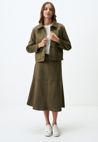 Olivgrüne Wildlederjacke mit Kragen und Fronttaschen, kombiniert mit einem passenden A-Linien-Rock, ergänzt durch weiße Sneakers.