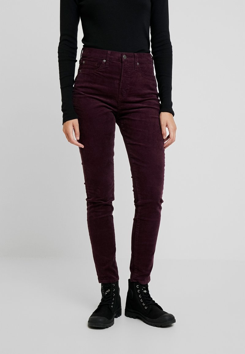 Personne portant un pantalon en velours côtelé slim fit violet foncé, un haut à manches longues noir et des bottines noires à lacets, debout sur un fond uni.