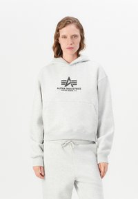 Donna che indossa un felpa con cappuccio grigio chiaro e pantaloni della tuta abbinati, in piedi davanti a uno sfondo bianco semplice.