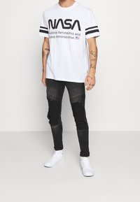 White cotton t-shirt featuring black "NASA" graphic and text, with black stripes on sleeves. Worn with distressed black jeans and white sneakers.