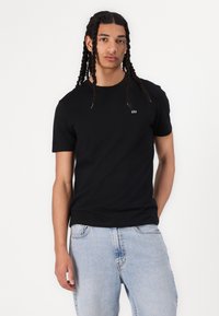 T-shirt en coton noir avec un col ras du cou, présentant un petit logo sur la poitrine. Associée à un jean en denim bleu clair. Texture lisse.