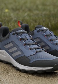 Paire de chaussures de trail Adidas bleu et noir avec semelles robustes sur un chemin de gravier, herbe floue en arrière-plan.