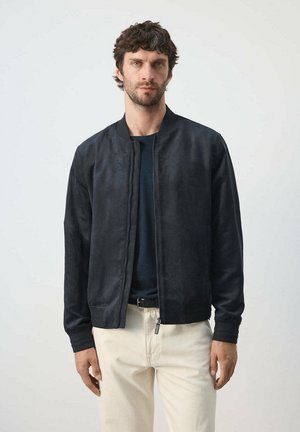 Hombre con cabello corto y oscuro y barba, vistiendo una chaqueta negra con cremallera, camisa oscura, pantalones crema y cinturón negro, de pie contra un fondo liso.