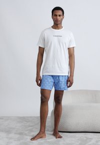 Λευκό βαμβακερό μπλουζάκι με μαύρο λογότυπο και μπλε σορτς με σχέδιο από μικρά λευκά σχήματα· το μοντέλο στέκεται ξυπόλυτο σε ανοιχτό γκρι επιφάνεια.