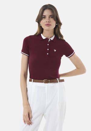 Bordoo polo särk valge krae ja äärega, millel on väike tikitud logo, koos kõrge vöökohaga valgete pükstega ja pruuni vööga.