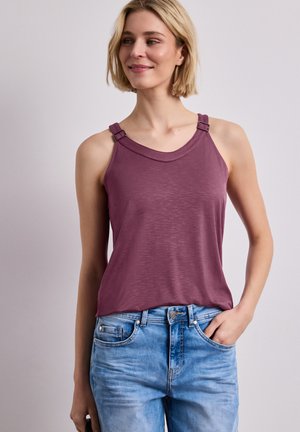 Kobieta ubrana w bezrękawnik o kolorze śliwkowym z regulowanymi ramiączkami i jasnoniebieskie jeansy, stoi z jedną ręką w kieszeni na neutralnym tle.