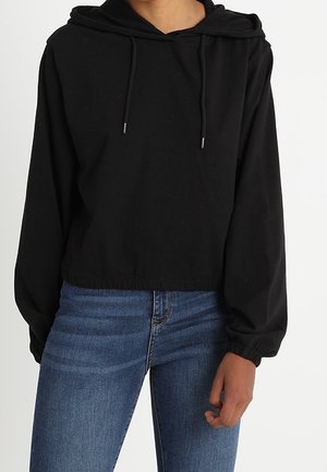 Persoon draagt een zwarte cropped hoodie met aantrekkoorden en blauwe spijkerbroek, staat met één hand ontspannen langs het lichaam.